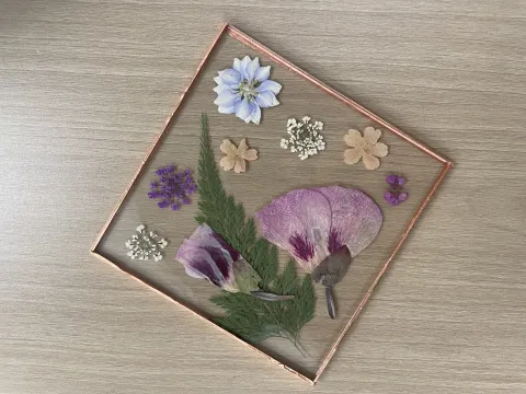 Clear coaster made from pressed flowers, purple, green, blue, yellow, and pink flowers pressed between 2 clear acrylic squares with rose gold washi tape taping the edges together to form a border.l