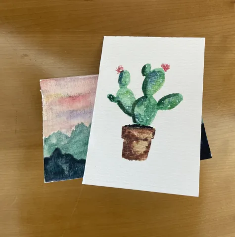 two greeting cards. one with a cactus planted in a brown pot with two red/pink blooms. The other one with a mountain landscape. green blue mountains with a sunset background.
