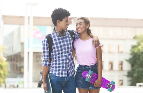 Two teens, one boy in a blue button up shirt and a girl in a pink shirt. They have their arm around each others shoulders walking. The boy has a backpack and they both have skateboards